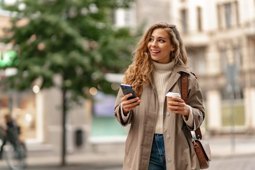 Young woman with curly blonde hair using the phone with a cup of coffee in hands on the city streets, portrait