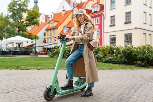 Young blonde woman riding a kick scooter in city street, full portrait