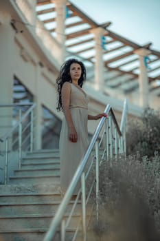 A woman in a dress stands on a set of stairs. The stairs are made of metal and are located in front of a building. The woman is wearing a necklace and a bracelet