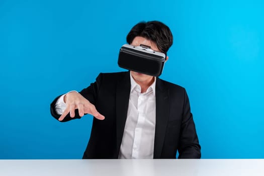 Asian businessman looking through VR connecting meta holding hologram object on table analyzing futuristic business metaverse innovation technology virtual reality isolate blue copyspace. Contrivance.