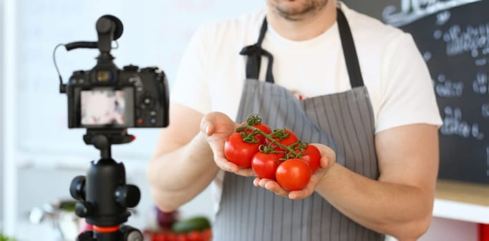 Vlogger Chef Showing Ripe Tomatoes Ingredient. Man in Apron Recording Big Red Vegetable on Camcorder for Culinary Vlog. Cooking Recipe. Male Hands Holding Fresh and Aromatic Food at Kitchen.