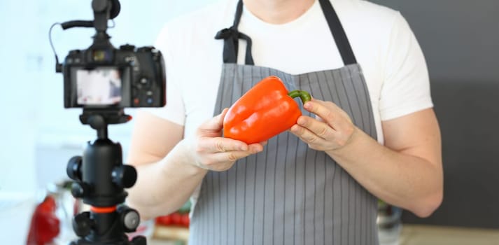 Chef Vlogger Showing Camcorder Orange Pepper. Man in Apron Recording Ripe Vegetable on Camera for Culinary Vlog in Kitchen at Home. Male Hands Holding Fresh Ingredient Horizontal Photography
