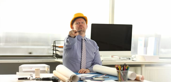 Serious Architect Working on Construction Outline. Caucasian Engineer Sitting at Office Workplace. Man Wear Formal Shirt, Tie and Yellow Helmet do House Drafting. Businessman Pointing at Camera