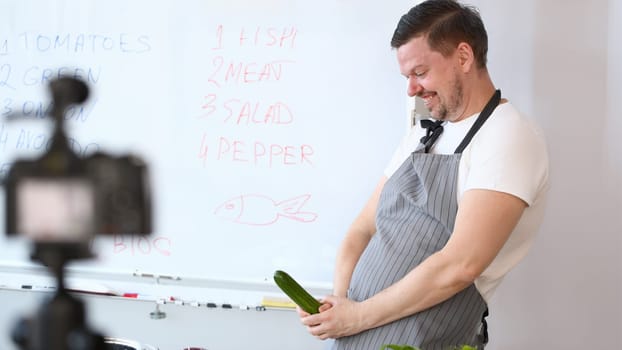 Smiling Vlogger Chef Recording Cucumber Joke. Cook in Apron Joking with Green Cuke on Camera. Culinary Vlog for Vegan at Home Kitchen. Cheerful Man Vegetable Ingredient for Cooking Vegetarian Food