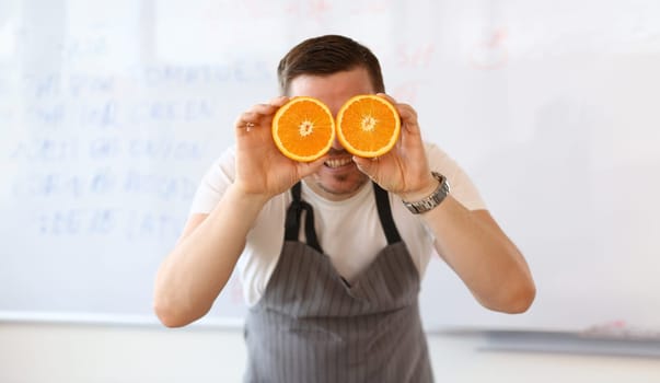 Vegan Chef Blogger Holding Cut Juicy Orange Fruit. Man with Colorful Citrus Halves on Eye in Kitchen on White Background. Concept of Healthy Food and Lifestyle. Smiling Person with Citrus in Hands