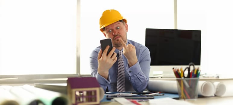 Architect Express Emotion at Engineering Office. Annoyed Caucasian Constructor in Shirt and Yellow Helm Giving Middle Finger to Mobile Phone in Hand. Angry Building Business Owner