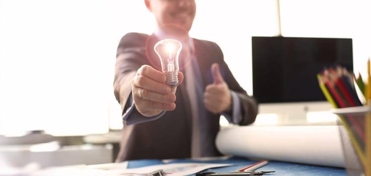 Inspiration Brand New Idea Originative Symbol. Happy Businessman Holding Traditional Light Bulb. Thumb Up Sign Manager in Suit with Lamp in Hand Sit at Table. Innovation Technology. Accounting Concept