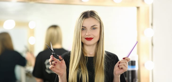 Hairdresser Woman Holding Scissors and Hairbrush. Blonde Hairstylist with Tool for Hair. Female Beautician with Comb for Hairdo Standing in Beauty Salon. Haircut Stylist Looking at Camera Shot