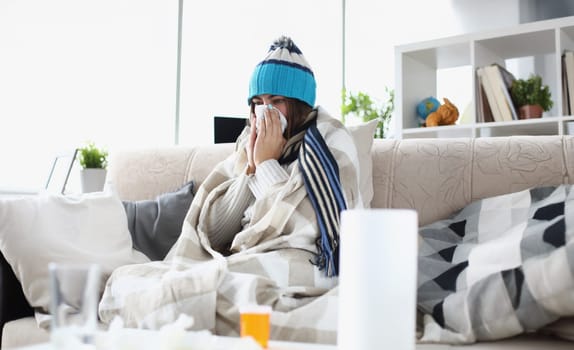Portrait of woman having severe flu at home indoors and sneezing into tissue. Girl sitting in living room covered in blanket and suffering from common and annoying cold. Hard virus concept