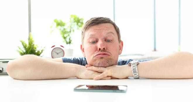 Man with funny facial expression staring at cellphone lying at table waiting for call concept