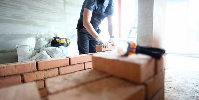 Male builder hand in gloves holding clay brick on background of brickwork wall. Repair and redevelopment apartments concept.