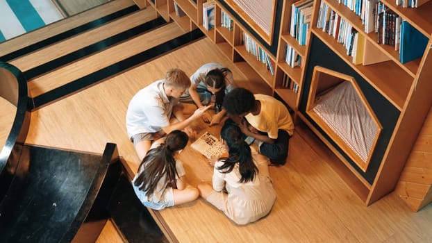 Top view of diverse children sitting in circle while looking at camera and waving hands at library surrounded with the stack of book. Multicultural highschool student greeting camera. Edification.