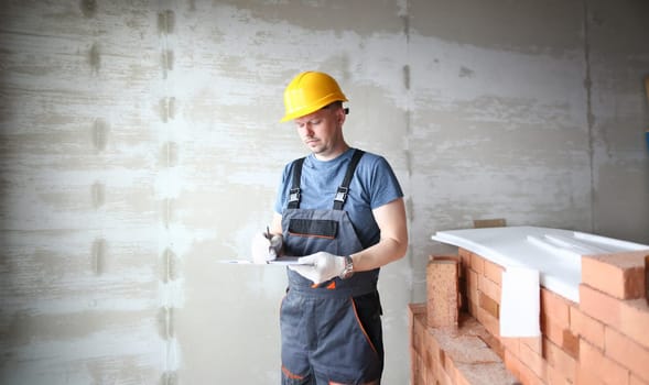 Male builder in yellow hard hat holds clipboard on background of an apartment under repair portrait. Estimated property value concept.