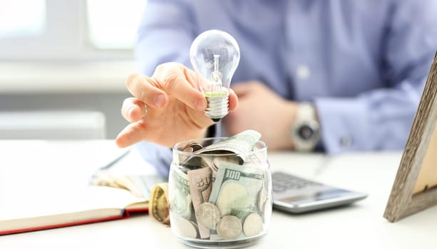 Male arm holding lightbulb over tip can full of US money closeup