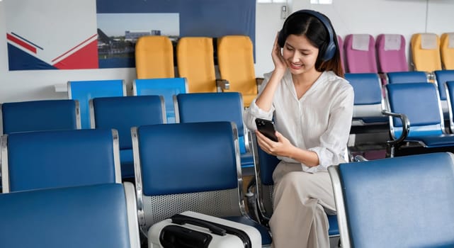 Asian woman listening to music on smartphone in airport waiting area. Concept of travel, technology, and relaxation.