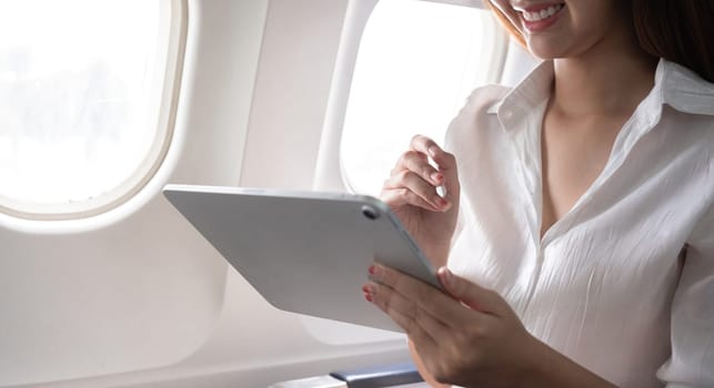 Asian woman using tablet on airplane. Concept of air travel, digital technology, and in-flight entertainment.