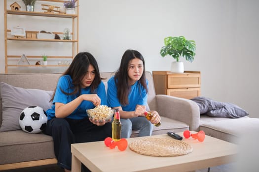 Asian female couple watching soccer game with snacks and drinks in living room. Concept of LGBTQIA sports fans and home entertainment.