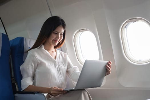 Asian woman working on laptop in airplane seat. Concept of air travel, remote work, and in-flight productivity.