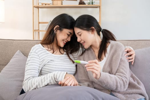 Asian lesbian couple celebrating positive pregnancy test at home. Concept of love, family planning, and LGBTQIA parenthood.