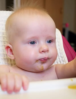 A cute baby with blue eyes has broccoli puree smeared all over their face. It looks like the baby is enjoying their meal while sitting at home.