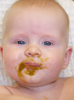 A cute baby with blue eyes has broccoli puree smeared all over their face. It looks like the baby is enjoying their meal while sitting at home.