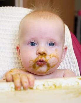 A cute baby with blue eyes has broccoli puree smeared all over their face. It looks like the baby is enjoying their meal while sitting at home.