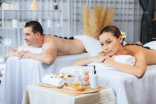 Hot stone massage at spa salon in luxury resort with day light serenity ambient, blissful couple customer enjoying spa basalt stone massage glide over body with soothing warmth. Quiescent