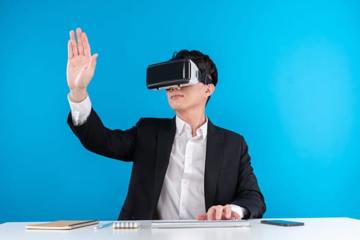 Asian businessman looking through VR connecting touchscreen hologram monitor in futuristic metaverse analyzing innovation technology business world virtual reality isolate blue copyspace. Contrivance.