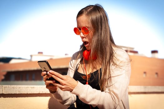 Young woman with cell phone in hands, writes messages and reads news online, using an app with a gesture of happiness.