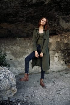 Mysterious woman in green trench coat stands on rocky hillside overlooking cave entrance