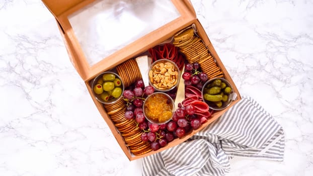 Flat lay. Hands are meticulously adding fresh red grapes to a bowl, complementing a beautifully arranged charcuterie box brimming with a variety of cheeses, olives, and cured meats, set against a sleek marble surface.