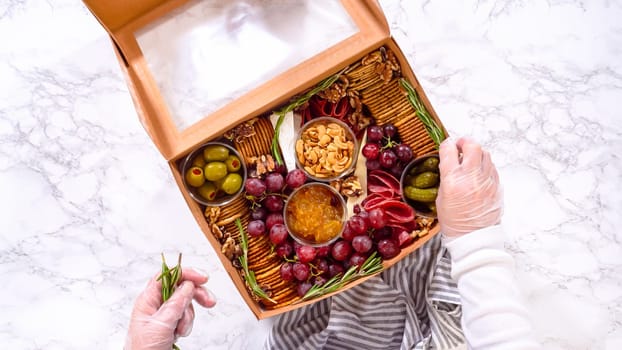 Flat lay. Hands are meticulously adding fresh red grapes to a bowl, complementing a beautifully arranged charcuterie box brimming with a variety of cheeses, olives, and cured meats, set against a sleek marble surface.
