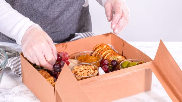 Hands are meticulously adding fresh red grapes to a bowl, complementing a beautifully arranged charcuterie box brimming with a variety of cheeses, olives, and cured meats, set against a sleek marble surface.