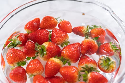 Ripe strawberries are submerged in water within a large glass mixing bowl, a step in washing the fruit to ensure cleanliness and longevity before storage or consumption.