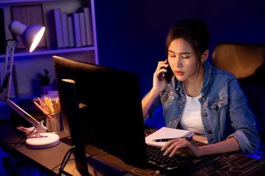 Young beautiful Asian creative woman calling on phone desk to customer or coworker explaining creator project's high profit while searching market analysis on pc at neon office at night. Stratagem.