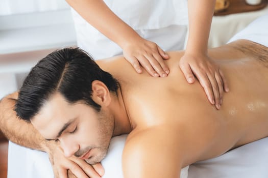 Caucasian man customer enjoying relaxing anti-stress spa massage and pampering with beauty skin recreation leisure in day light ambient salon spa at luxury resort or hotel. Quiescent