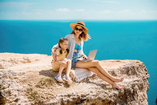 A woman and a child are sitting on a rock overlooking the ocean. The woman is using a laptop while the child looks on. Concept of relaxation and bonding between the two