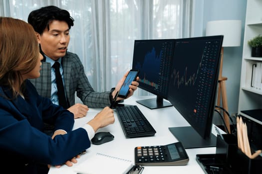 Smiling businessman showing dynamic stock market data on mobile phone to woman writing to memo for analyzing profit value currency rate online website program application at home office. Infobahn.