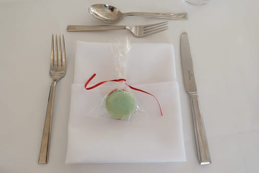 A table setting with a white napkin, silverware, and a green macaron wrapped in plastic with a red ribbon.