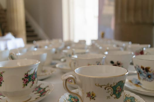 A close-up of a table set with multiple ornate porcelain teacups and saucers, each with intricate floral designs. The background is softly blurred, highlighting the teacups in the foreground.