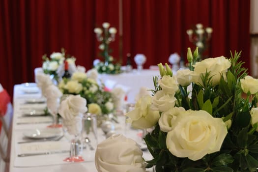 A beautifully decorated wedding reception table with white roses and elegant table settings. The background features red curtains and additional floral arrangements.