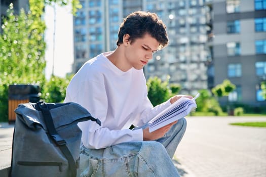 Handsome young male university college student reading a book sitting on outdoor steps. Education, knowledge, youth 19-20 years old, literary hobby and leisure