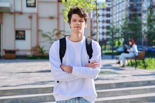 Portrait of confident smiling college student guy, young male with crossed arms with backpack looking at camera outdoor near educational building. Education, training, 19,20 years age youth concept