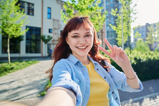 Selfie portrait of young beautiful attractive happy red-haired female looking at camera outdoor. Sunny day teenage girl student 19-20 years old hand showing victory peace gesture educational building