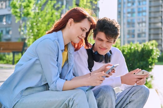 Teenage youth friends guy and girl university college students sitting outdoor on campus steps talking laughing using smartphone. Technology, lifestyle concept