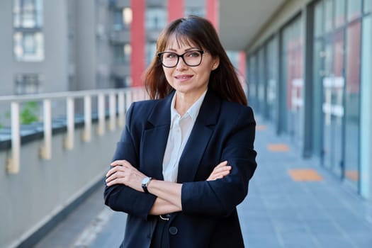 Mature confident successful business woman with crossed arms in black suit looking at camera outdoors, backdrop of modern city. Business, entrepreneurship mentoring insurance sales advertising work