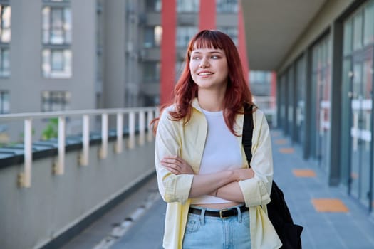 Urban portrait of young confident red-haired female with backpack with crossed arms, city outdoor. Lifestyle, youth 19-20 years old concept