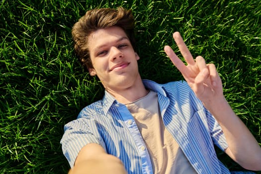 Top view, happy handsome smiling young male looking at camera, lying on background of green lawn grass, showing hand gesture peace victory success