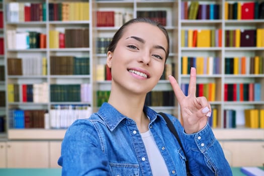 Selfie photo portrait of smiling cool high school student girl showing hand gesture for victory success, inside library of educational building. Education, adolescence, lifestyle concept