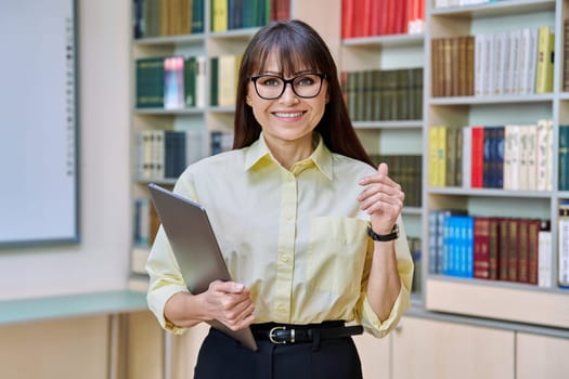 Middle aged elegant woman holding laptop computer inside library. Positive smiling business female teacher professor, leader, manager, mentor, psychologist, counselor, social worker at workplace